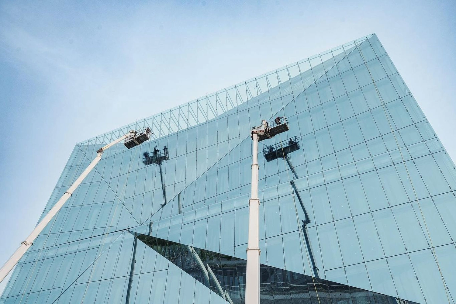 Break Facility Service - Zwei Hubarbeitsbühnen reinigen unter klarem Himmel die Glasfassade eines modernen blauen Gebäudes.