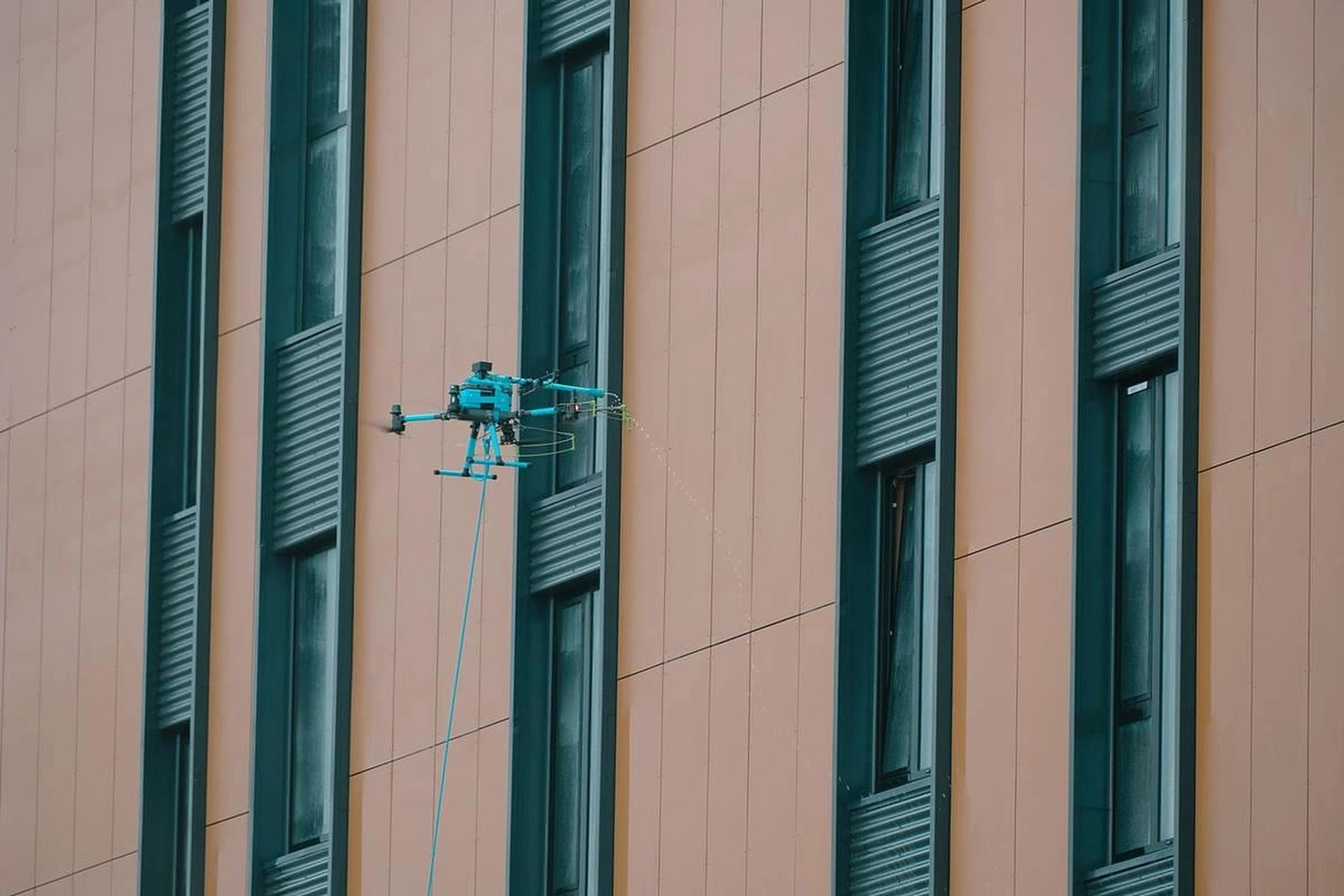 Break Facility Service - Eine blaue Drohne besprüht an einer Leine befestigt eine Gebäudefassade.