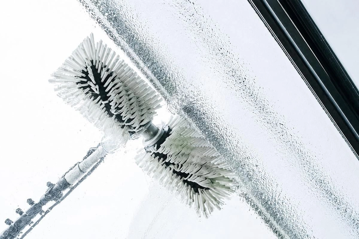 Break Facility Service - Eine rotierende, weiße, zylindrische Bürste reinigt eine transparente Glasoberfläche und hinterlässt dabei eine saubere Spur durch die Wassertropfen.
